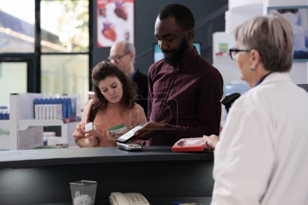 Foto de Young adult client putting smartphone on pos terminal paying for supplements in pharmacy after discussing health care treatment with pharmacist. Client at drugstore counter buying prescription pills - Imagen libre de derechos
