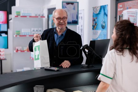 Téléchargez les photos : Un employé du drugstore a remis un sac en papier rempli de médicaments à un client âgé après l'avoir aidé avec une prescription médicale. Clients âgés achetant des pilules, vitamines, suppléments en pharmacie - en image libre de droit