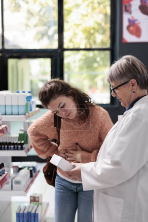 Téléchargez les photos : Drugstore employee helping sick customer with abdominal cramps offering stomachache painkillers while working in pharmacy. Client having gastritis looking for pills to cure food poisoning - en image libre de droit