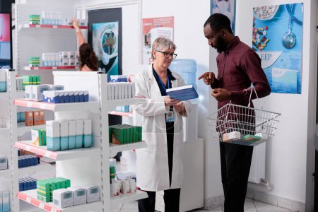 Téléchargez les photos : African american client holding box of pills asking for help to elderly pharmacist, drugstore worker explaining drugs medical leaflet. In background customers doing health care shopping - en image libre de droit