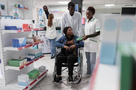 Téléchargez les photos : Pharmacienne expliquant femme afro-américaine en fauteuil roulant suppléments instruction en pharmacie. Client ayant une déficience physique et travailleur social acheter des vitamines, toute l'équipe noire - en image libre de droit