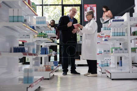 Photo for Senior drugstore employee helping customer with pharmaceutical products trying to find cardiology drugs in pharmacy. Client looking at medicaments on drugstore shelves. Medicine service and concept - Royalty Free Image