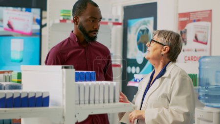 Téléchargez les photos : African american man asking senior woman for medical support, looking to buy bottle of pills from shelves. Pharmacist explaining disease treatment on prescription to young adult. - en image libre de droit