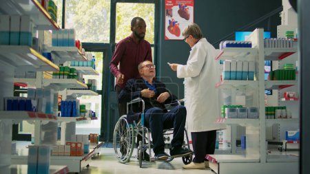 Photo for Social caretaker helping elderly person in wheelchair to come at drugstore, talking to pharmacist about fever measurement with thermometer. Client with physical disability and caregiver. Tripod shot. - Royalty Free Image