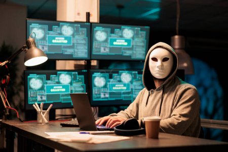 Foto de Hacker usando máscara y capucha para hackear el sistema informático, irrumpiendo en los servidores de la compañía para robar big data. Hombre enmascarado buscando peligroso y aterrador, impostor creando malware de seguridad. - Imagen libre de derechos
