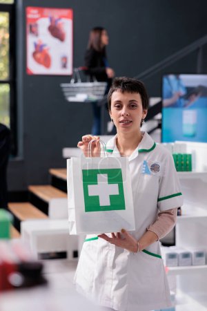 Téléchargez les photos : Femme pharmacienne vendant des médicaments et tenant portrait sac à provisions en papier. Consultant pharmaceutique aider à choisir des pilules, donner l'achat de médicaments et regarder la caméra - en image libre de droit