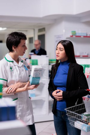 Téléchargez les photos : Drugstore worker giving customer assistance in choosing food poisoning remedy. Young asian woman holding hand on stomach and explaining abdominal pain symptom to pharmacy employee - en image libre de droit