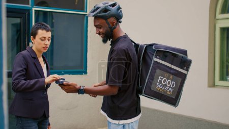 Photo for Smiling woman paying for food delivery service with credit card, pos contactless payment at office front door. Restaurant courier with backpack holding terminal, giving fastfood meal order. - Royalty Free Image