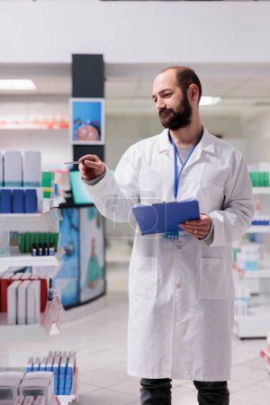 Foto de Drugstore employee holding clipboard while checking pills packages writing medication name on papers, working in pharmacy. Pharmacist is responsible for maintaining the inventory of drugs - Imagen libre de derechos