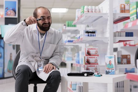Foto de Pharmacist standing on chair in empty pharmacy holding medical otoscope looking at camera while waiting for clients. Health care facility equipped with pharmaceutical products and pills - Imagen libre de derechos