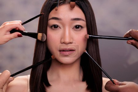 Photo for Fashion model getting make up in professional studio and looking at camera. Makeup artists team hands holding brushes around young adult attractive asian woman close up portrait - Royalty Free Image