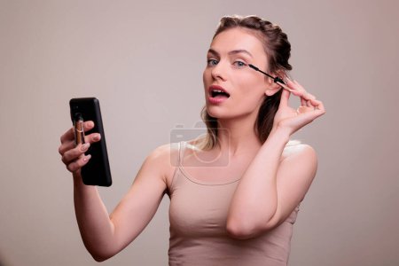 Téléchargez les photos : Femme appliquant le mascara sur les cils et regardant l'écran du smartphone. Belle jeune femme faisant naturel quotidien maquillage avec bouche ouverte et en utilisant la caméra avant de téléphone mobile comme miroir - en image libre de droit