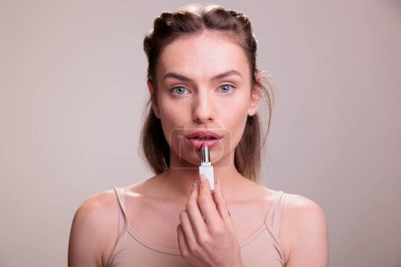 Foto de Mujer joven aplicando lápiz labial rosa y haciendo retrato de maquillaje desnudo. Modelo de producto cosmético hermoso natural usando el tubo del lipbalm del color, mostrando la rutina del maquillaje de los labios y mirando la cámara - Imagen libre de derechos