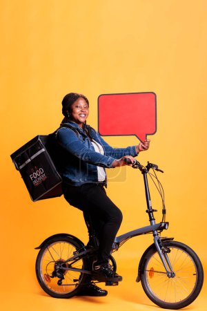 Photo for Pizzeria courier holding red speech bubble advertising takeaway service while riding bicycle preparing to deliver takeout order to customers. Courier standing in studio with yellow background - Royalty Free Image