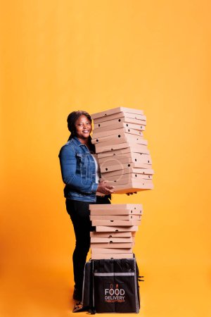 Foto de Repartidora sonriente sosteniendo una pila llena de cajas de pizza preparándose para entregar a los clientes durante la hora del almuerzo. Restaurante Emloyee con comida para llevar mochila térmica entrega comida para llevar - Imagen libre de derechos