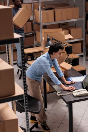 Photo for Warehouse manager checking shipping details on laptop computer before start packing customers orders, putting clothes in carton boxes, preparing for delivery. Woman working in delivery department - Royalty Free Image