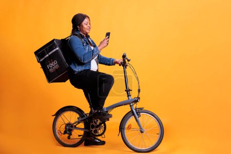 Téléchargez les photos : Femmes afro-américainestrès debout sur le vélo en studio avec fond jaune tout en vérifiant adreess client au téléphone avant de commencer la livraison repas à emporter. Pizzeria courier portant sac à dos à emporter - en image libre de droit