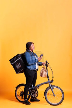 Photo for Deliverywoman holding smartphone checking client adreess on fast food app before start delivering lunch orders. Restaurant employee carrying takeout backpack while riding bike as transportation - Royalty Free Image