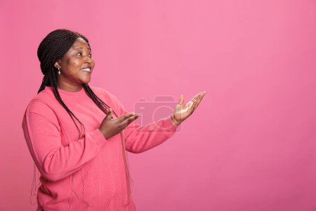 Photo for Portrait of smiling woman advertising promotional product in studio, posing for advertisement over pink background. Confident model with stylish hairstyle looking at camera during ad shoot - Royalty Free Image