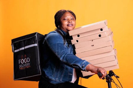 Téléchargez les photos : Pizzeria livreur portant pile de carton plein avec pizza livrer commande de nourriture à emporter au client pendant l'heure du déjeuner, debout en studio. Employé du restaurant utilisant le vélo comme moyen de transport - en image libre de droit