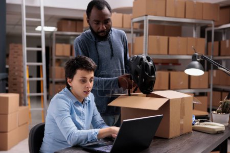 Photo for Storehouse employee putting customer helmet order in carton box preparing delivery for client while discussing transportation details with manager. Diverse team working in warehouse - Royalty Free Image