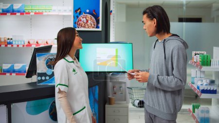 Foto de Asian pharmacist showing box of pills to young man holding prescription paper, specialist explaining treatment in pharmacy store. Consultant helping client with medical products. - Imagen libre de derechos