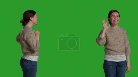 Foto de Close up of young adult greeting person with wave, smiling in studio. Relaxed casual woman waving hi or hello on camera, standing over full body green screen background and saying goodbye. - Imagen libre de derechos