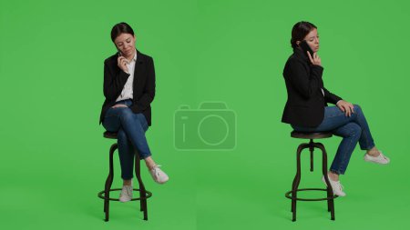 Photo for Caucasian businesswoman answering call on smartphone, sitting on chair in studio background. Office worker talking on remote telephone line using mobile phone, full body green screen backdrop. - Royalty Free Image