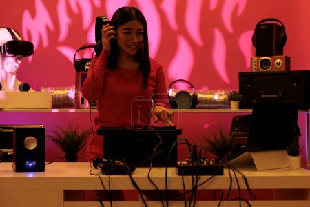 Téléchargez les photos : Musicien asiatique avec écouteurs mélangeant sons électroniques et techno jouant de la musique devant la foule en utilisant la console de mixage en studio. Artiste avec chemisier rose appréciant de chanter en club la nuit - en image libre de droit