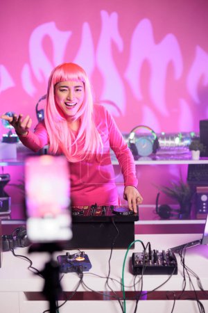 Photo for Artist dancing in studio while mixing music at professional mixer console filming music session with mobile phone camera. Dj with pink hair performing electronic concert having fun at night in club - Royalty Free Image