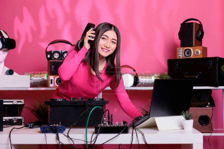 Téléchargez les photos : Musicien asiatique portant un casque lors de l'exécution de la musique électronique en utilisant des platines professionnelles en studio sur fond rose. Dj lecture de sons stéréo avec électronique et microphone - en image libre de droit