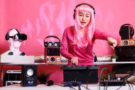Photo for Asian performer wearing headphones while playing electronic music using professional mixer console, enjoying performing in night club. Musician with pink hair standing at dj table mixing sounds - Royalty Free Image