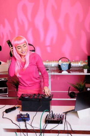 Photo for Artist listening techno music at headphones while mixing song at professional turntables, having fun with fans in club at night. Asian woman with pink hair doing performance with audio equipment - Royalty Free Image