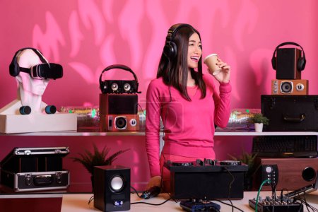 Photo for Smiling artist playing techno sound at professional turntables while drinking coffee, enjoying playing songs at night in club. Asian performer creating musical performance with remix music - Royalty Free Image