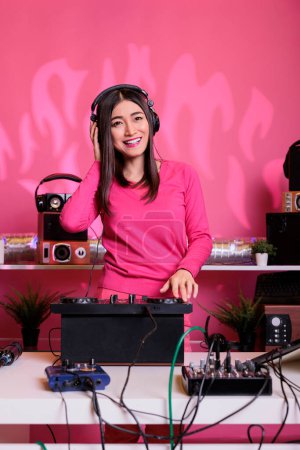 Photo for Artist standing at dj table mixing electronic sounds with techno using professional mixer console, having fun in studio over pink background. Asian musician playing stereo sounds with electronics - Royalty Free Image