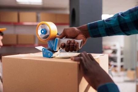 Photo for African american man hands holding packing tape gun and sealing cardboard box. Warehouse worker closing parcel with adhesive and preparing customer order for dispatching close up - Royalty Free Image