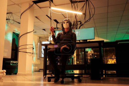 Foto de Hacker con máscara mirando la imagen holográfica en realidad aumentada, trabajando con holograma para hackear la red informática. Joven ladrón anónimo disfrutando de inteligencia artificial por la noche. - Imagen libre de derechos
