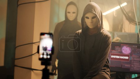 Young web spy recording ransomware video to receive reward, stealing information and broadcasting live threat. Dangerous hacker with anonymous mask doing illegal activity. Handheld shot.