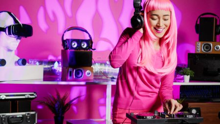 Téléchargez les photos : Asian artist using cutting-edge technology to remix music, listening song using headphones while performing in club at night time. Musician standing at dj table playing with professional mixer - en image libre de droit