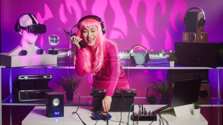 Téléchargez les photos : Musicien asiatique debout à la table de DJ jouant de la musique techno en utilisant une console de mixage professionnelle, appréciant de jouer la nuit en club. Interprète joyeux s'amuser avec les fans pendant le concert techno - en image libre de droit
