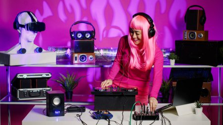 Foto de Cheerful performer listening music into headphones while mixing techno sound using professional mixer console. Artist standing at dj table dancing and having fun, enjoying party in night club - Imagen libre de derechos