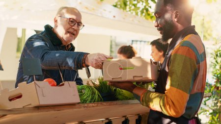 Photo for African american vendor selling fresh homegrown products at outdoors greenmarket, talking to senior customer about healthy nutrition. Farmer showing organic produce. Handheld shot. - Royalty Free Image