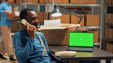 Téléchargez les photos : L'homme afro-américain parle au téléphone fixe et utilise un écran vert sur un ordinateur portable, travaillant avec un écran chromatique isolé à l'entrepôt. Storehouse propriétaire avec téléphone et maquette. - en image libre de droit