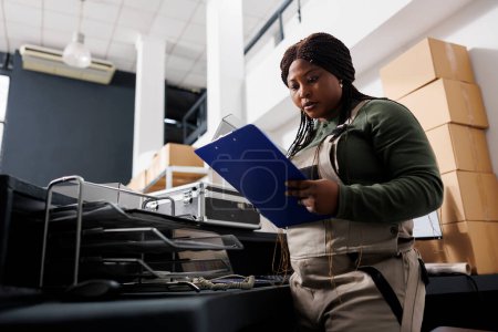 Photo for Employee holding clipboard analyzing goods logistics, working at customers orders preparing packages for delivery in warehouse. African american stockroom worker working at merchandise inventory - Royalty Free Image