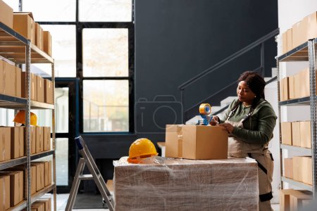Photo for African american employee preparing customers order, using adhesive tape to pack products in cardboard boxes in storage room. Small business worker wearing industrial overall working in storehouse - Royalty Free Image
