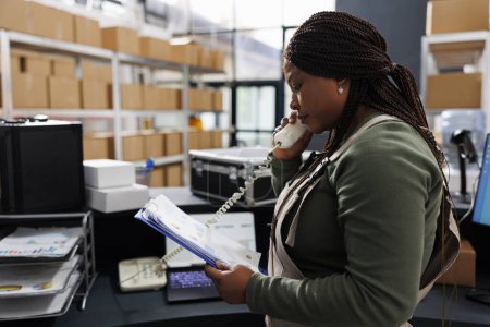 Photo for African american worker talking at landline phone with remote customer discussing order details. Storehouse employee holding clipboard, analyzing goods inventory report in storage room. - Royalty Free Image
