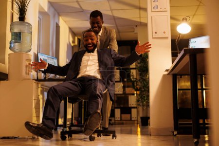 Photo for Smiling happy workers riding desk chair during sports race competition during break time in modern startup office. Cheerful african american managers having fun, enjoying doing moving activity - Royalty Free Image