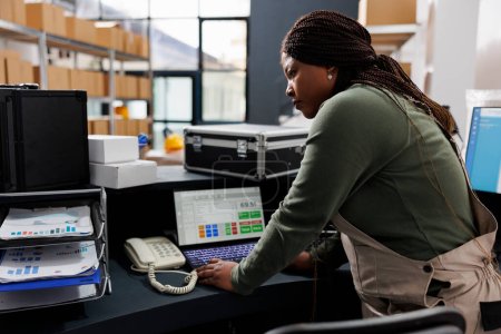 Téléchargez les photos : Superviseur afro-américain analysant la logistique des marchandises sur ordinateur portable, travaillant au rapport d'inventaire des marchandises. Employé d'entrepôt portant l'ensemble industriel travaillant aux commandes des clients - en image libre de droit
