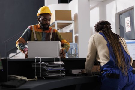 Photo for African american package handler putting packed parcel for shipment on checkout desk. Warehouse worker wearing protective helmet carrying heavy cardboard box to storage room reception - Royalty Free Image