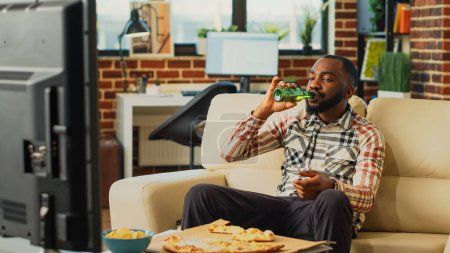 Photo for Young male adult eating slice of pizza at tv, laughing in living room on couch. Happy relaxed person enjoying takeaway meal and binge watching favorite tv show, having fun at home. - Royalty Free Image